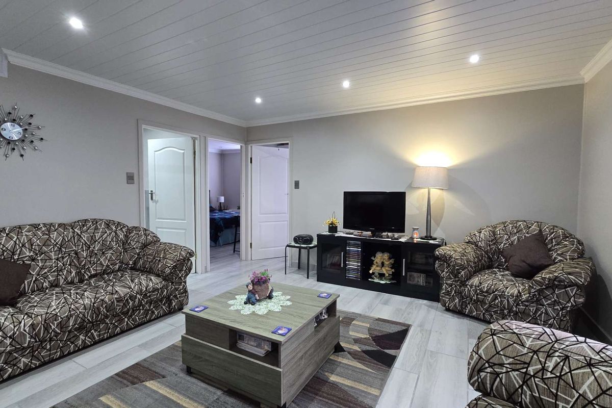 A well-maintained living room featuring light-toned wood-look flooring, recessed ceiling lighting, and a clean, neutral wall finish with access to adjacent rooms.