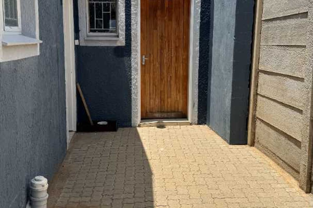 A paved side exterior area featuring a wooden door, security window grille, and textured dark blue exterior wall finish.