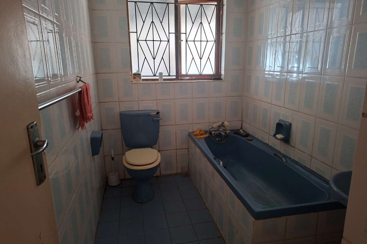 A dated bathroom featuring blue sanitaryware, including a built-in bathtub and toilet, with fully tiled walls and floors and a window with security bars.