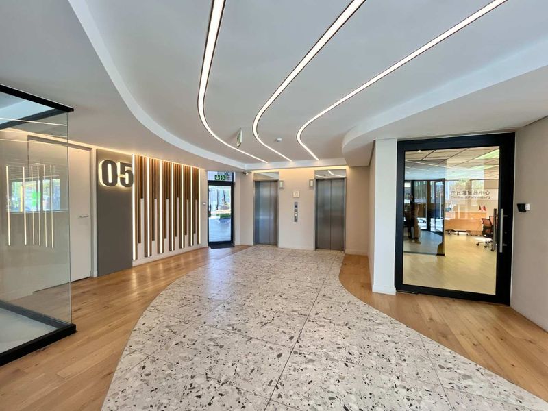Modern office building lobby featuring high-end finishes, including a combination of terrazzo and hardwood flooring, recessed linear ceiling lights, and stainless steel elevator access.