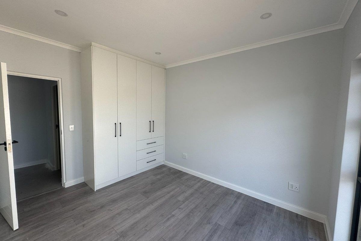 A bright, modern bedroom featuring neutral grey walls, wood-look laminate flooring, and built-in white cabinetry with integrated drawers.