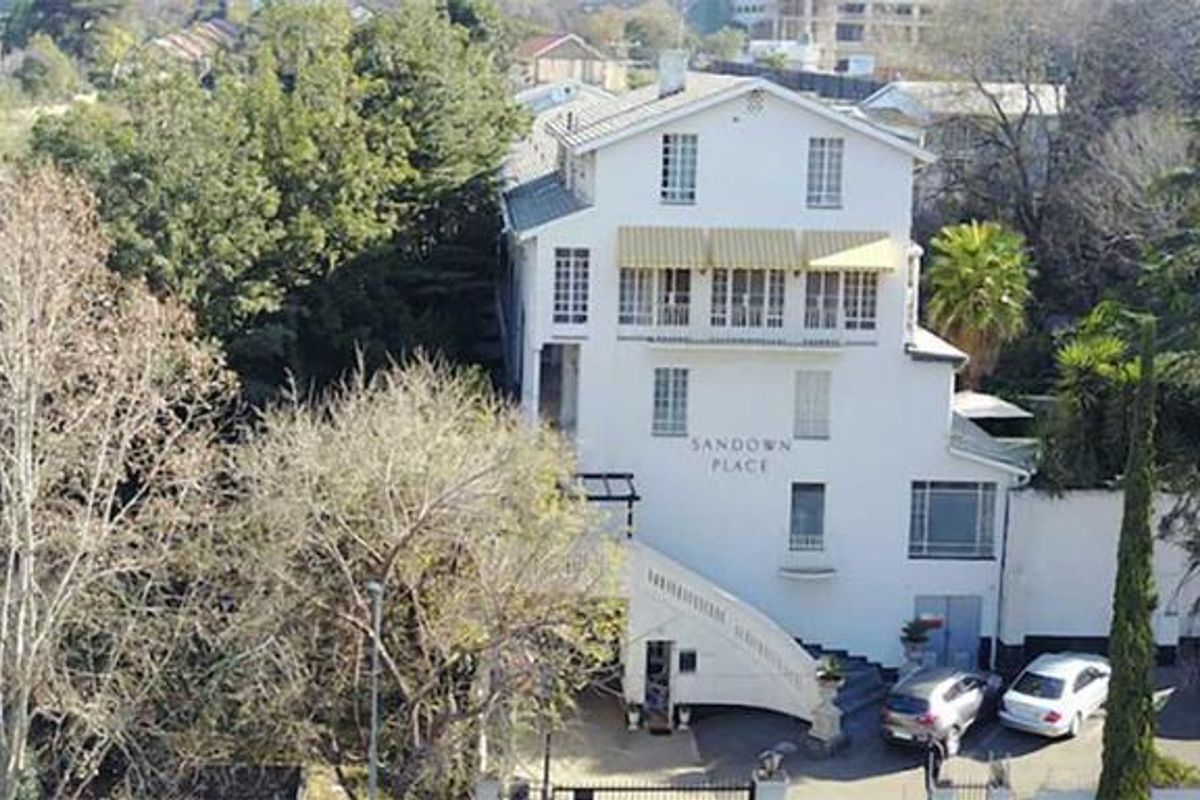 Aerial view of Sandown Place, a multi-story apartment building featuring an external staircase, pitched roof, and dedicated parking area surrounded by mature trees.
