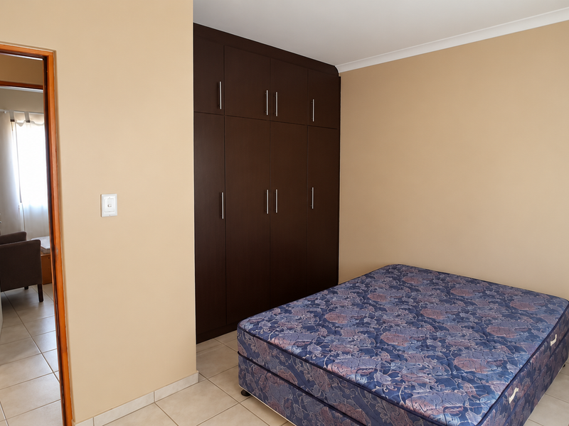 A bedroom featuring dark wood built-in cupboards, neutral wall paint, and tiled flooring, with an adjacent view into a bathroom area.