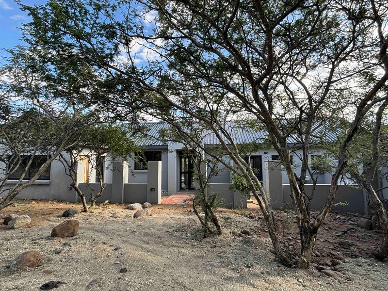 Front elevation of a single-story house featuring a corrugated metal roof, masonry boundary walls, and a paved entrance path surrounded by natural landscaping.