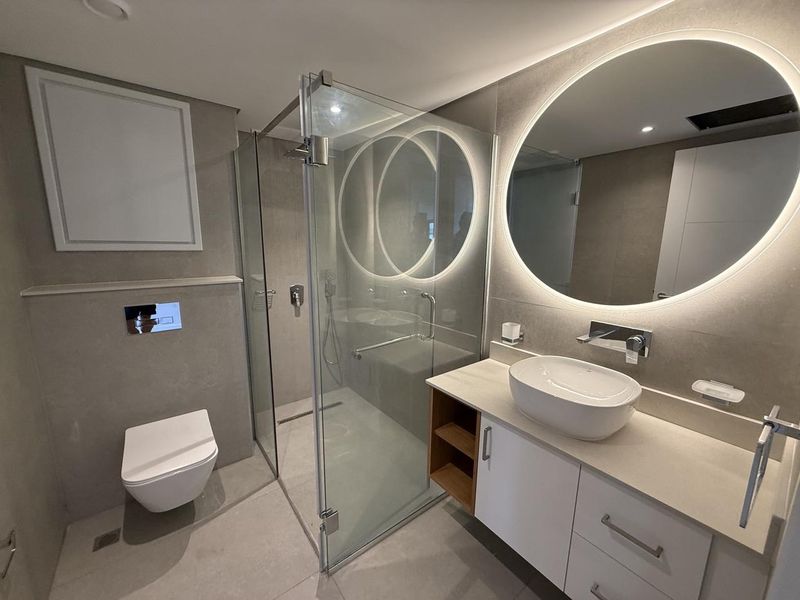 Modern bathroom featuring a wall-mounted toilet, glass-enclosed walk-in shower, and a contemporary vanity with a vessel sink and backlit circular mirror.