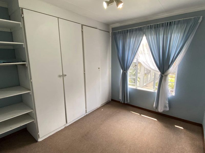 A bright bedroom featuring built-in cupboards, integrated shelving, and a window with security bars, offering a functional and clean living space.