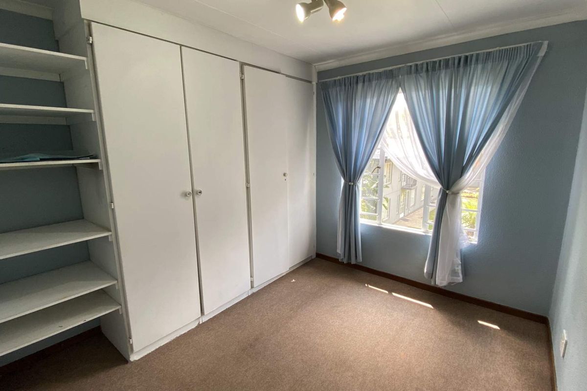 A bright bedroom featuring built-in cupboards, integrated shelving, and a window with security bars, offering a functional and clean living space.
