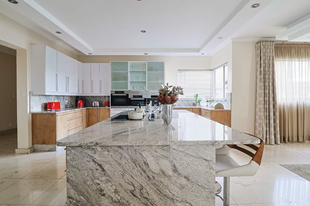 Modern kitchen featuring a large granite-topped island, built-in stainless steel ovens, glass-fronted cabinetry, and polished tile flooring.