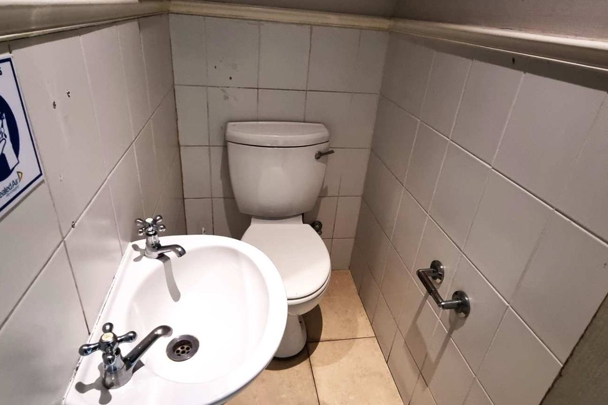 A compact office restroom featuring a wall-mounted basin with dual taps and a standard toilet, finished with white wall tiling.