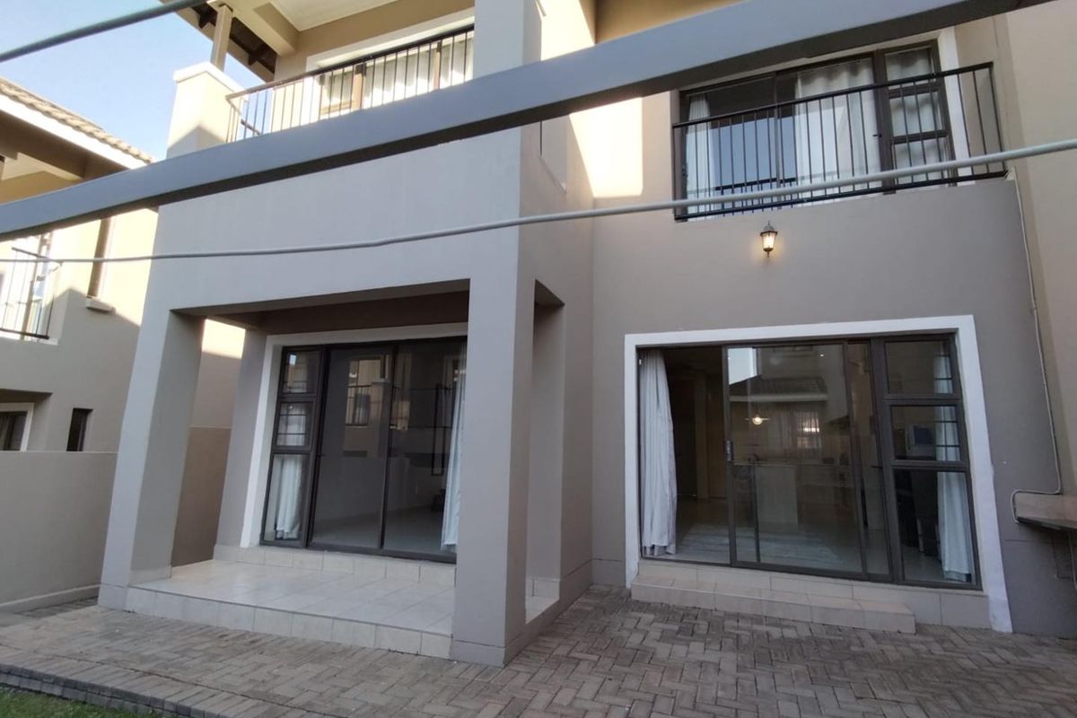 Rear view of a modern townhouse featuring a paved patio area, sliding glass doors, and an upper-level balcony with metal railings.