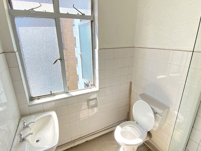 A compact bathroom featuring white wall tiles, a pedestal basin, toilet, and a glass shower screen, requiring cosmetic updates to improve its condition.