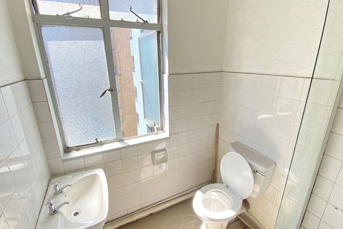 A compact bathroom featuring white wall tiles, a pedestal basin, toilet, and a glass shower screen, requiring cosmetic updates to improve its condition.