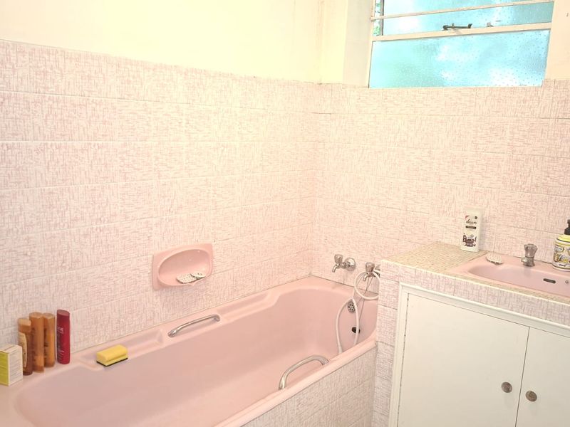 A dated bathroom featuring a pink built-in bathtub, integrated vanity sink, and textured wall tiling with a louvered window.
