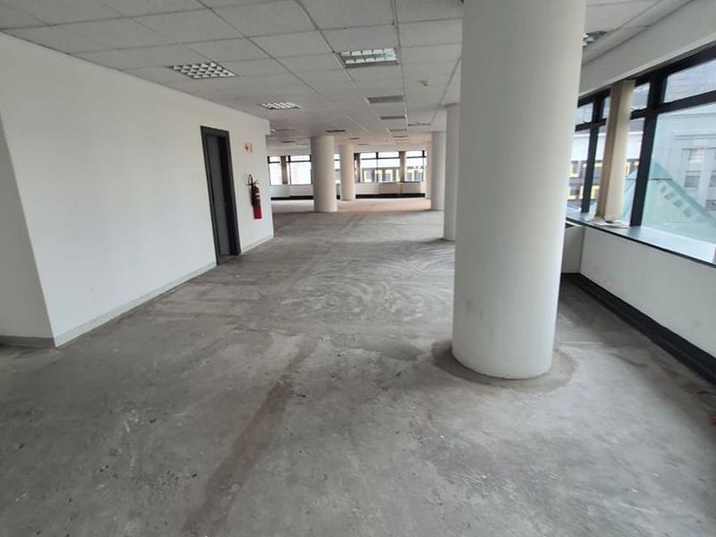 An expansive, open-plan office space featuring structural columns, large windows for natural light, and a suspended ceiling, currently awaiting floor finishing.