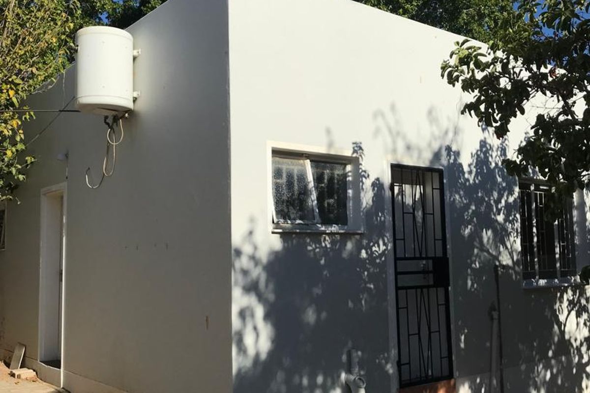 Exterior view of a small outbuilding featuring a white rendered facade, security gate, tiled entrance steps, and an externally mounted electric geyser.