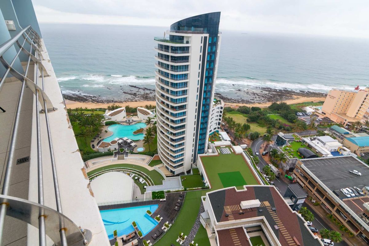 High-rise aerial view overlooking the coastline, featuring modern apartment architecture, landscaped grounds, and swimming pool facilities.