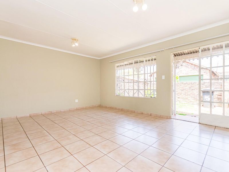A bright, empty living room featuring neutral tiled flooring, security bars on the window, and direct access to the rear exterior area.
