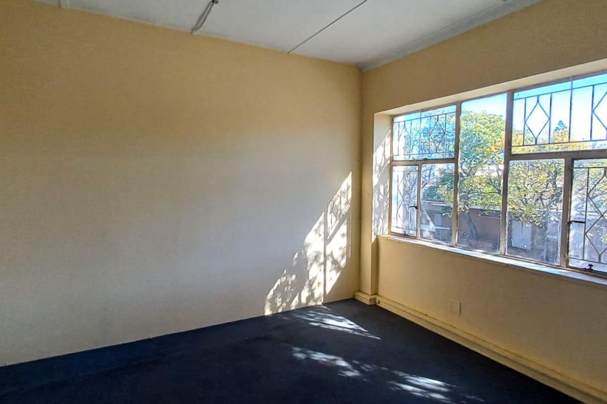 An empty room featuring large windows with security bars, dark carpeting, and neutral wall finishes, suitable for office or residential use.