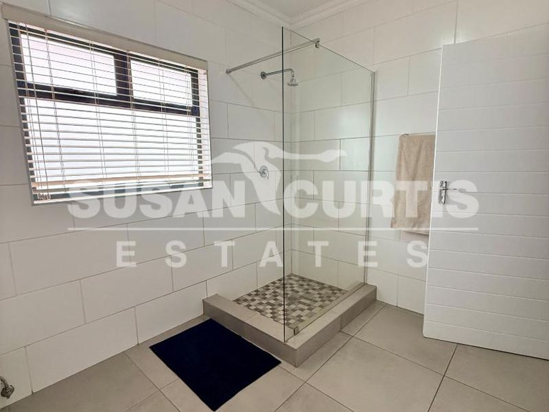 A clean, modern bathroom featuring a walk-in shower with a glass screen, neutral wall and floor tiling, and a window with blinds.