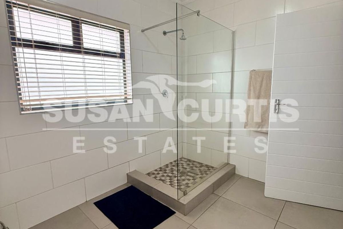A clean, modern bathroom featuring a walk-in shower with a glass screen, neutral wall and floor tiling, and a window with blinds.