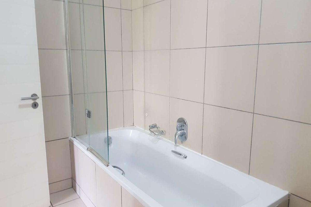 A clean, functional bathroom featuring a built-in bathtub with a glass shower screen and neutral wall tiling.