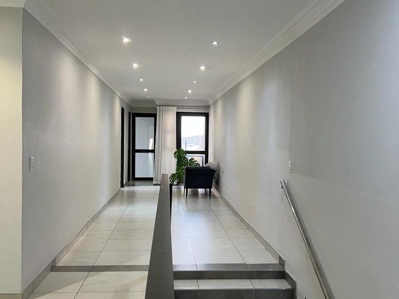 A bright, modern hallway featuring tiled flooring, recessed downlighting, and a staircase with a sleek metal handrail leading to a glass-paneled door.