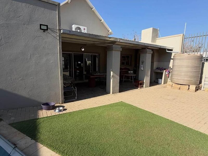 Rear exterior view featuring a covered patio, paved courtyard, artificial lawn, and a swimming pool with an integrated water storage tank.