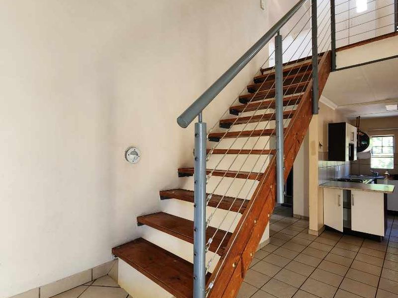 An open-plan hallway featuring a timber staircase with metal balustrades, ceramic tiled flooring, and a vaulted ceiling leading to an adjacent kitchen area.