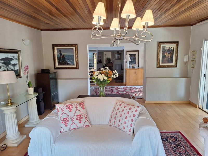 A well-maintained living room featuring a warm timber tongue and groove ceiling, hardwood flooring, and a classic chandelier, leading into an adjacent living space.