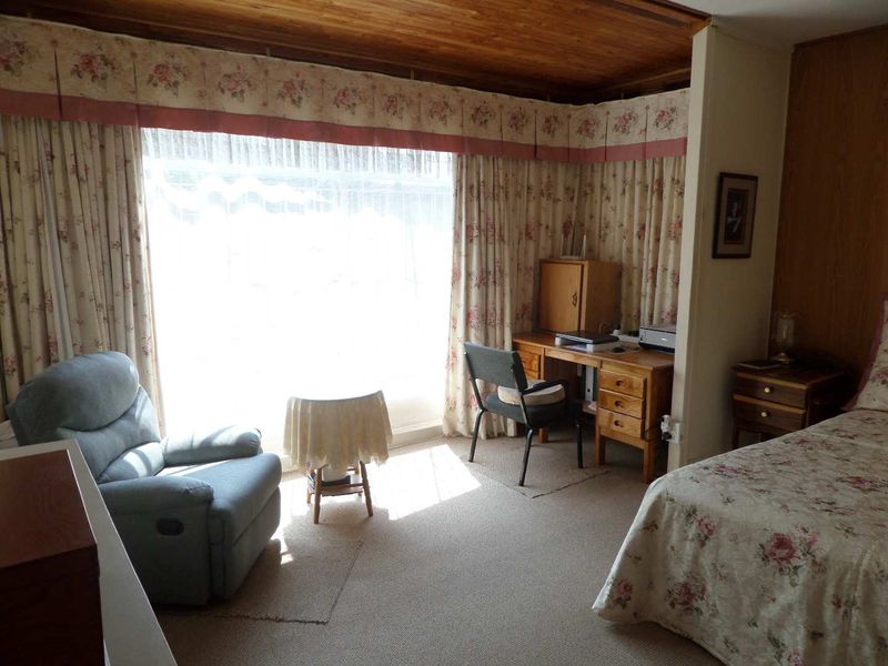 A bedroom featuring a timber ceiling, carpeted flooring, and a large window with floral curtains, offering a dated but functional living space.