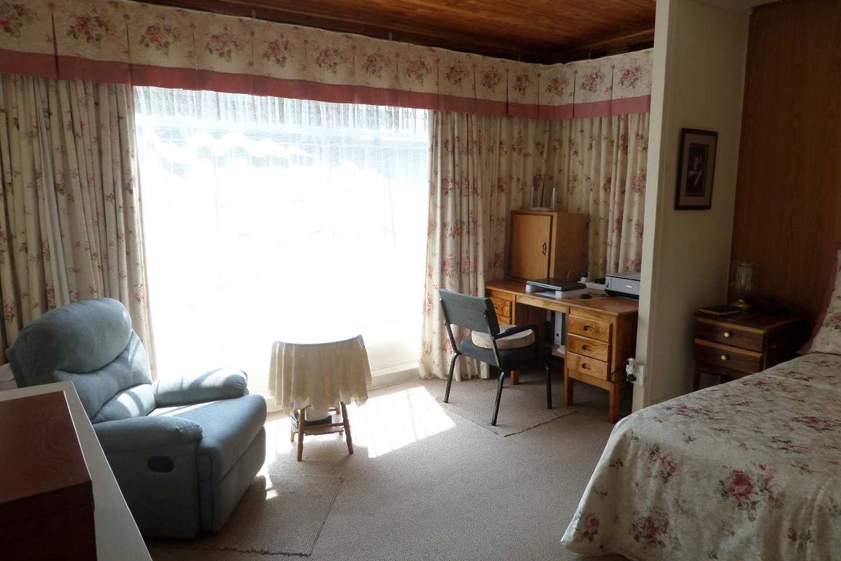 A bedroom featuring a timber ceiling, carpeted flooring, and a large window with floral curtains, offering a dated but functional living space.