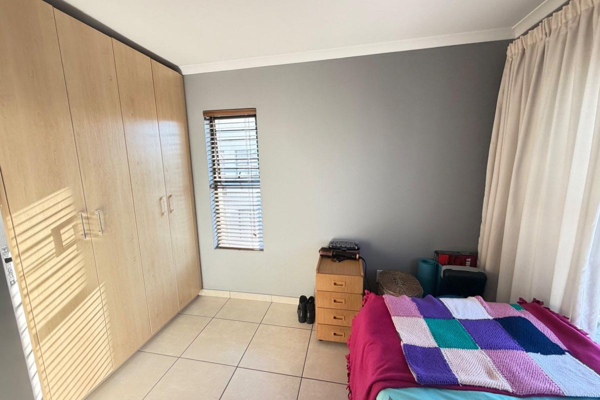 A bedroom featuring light wood-finish built-in cupboards, tiled flooring, and a window with fitted blinds.
