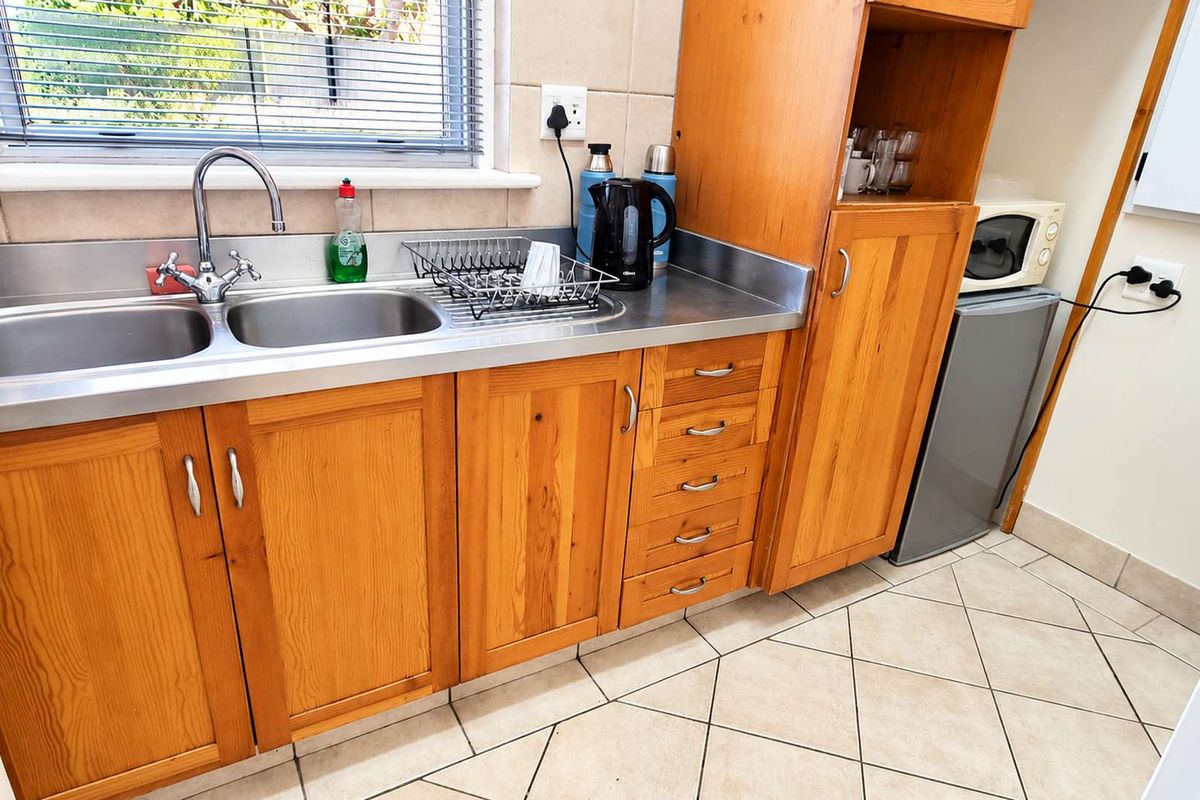 A functional office kitchenette featuring wooden cabinetry, a stainless steel double sink, tiled backsplash, and durable floor tiles.