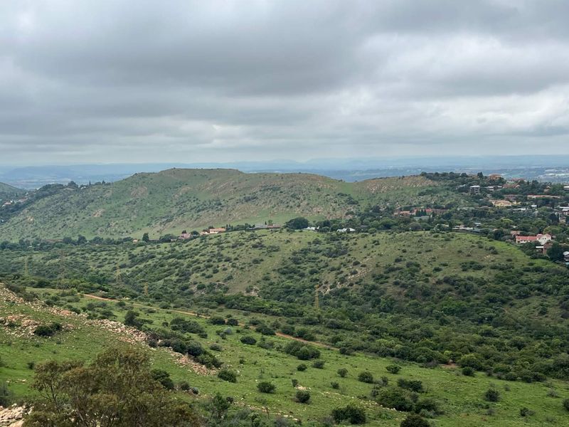 A scenic elevated view overlooking a lush green hilly landscape with distant residential areas under a cloudy sky.