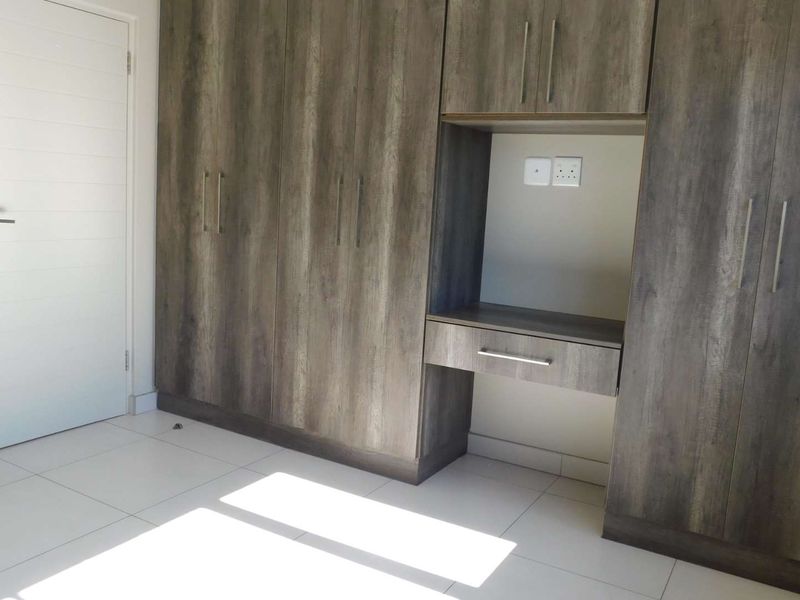 A bright bedroom featuring modern wood-finish built-in cupboards with an integrated desk area and clean tiled flooring.