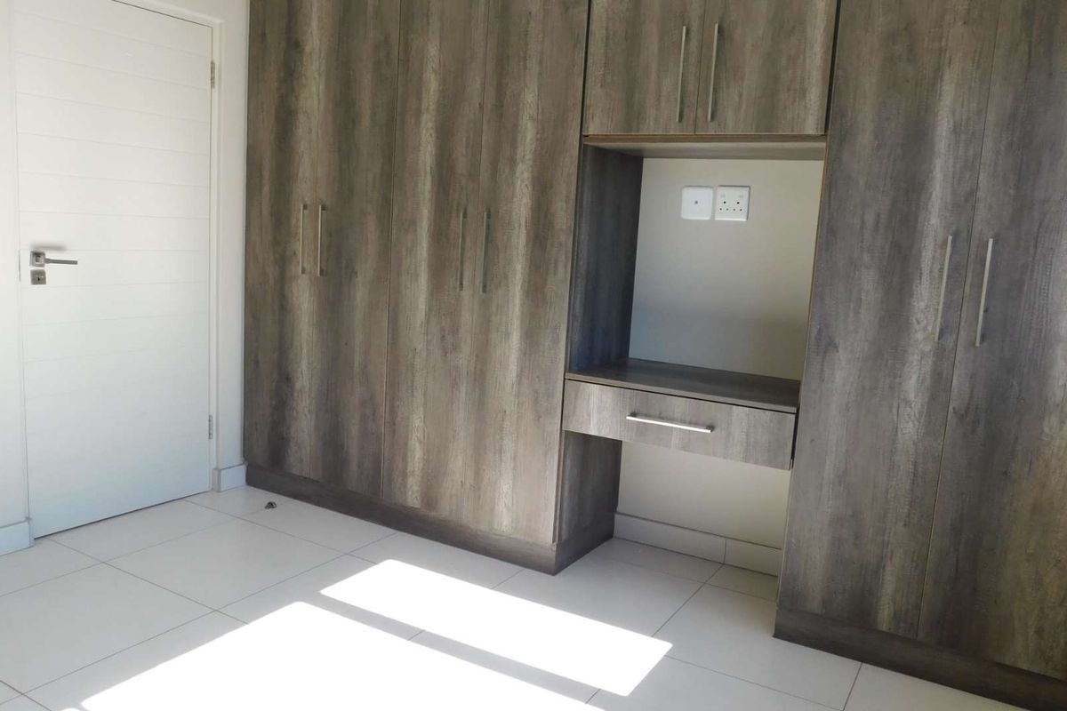 A bright bedroom featuring modern wood-finish built-in cupboards with an integrated desk area and clean tiled flooring.