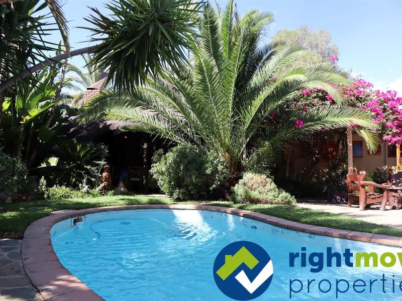A well-maintained outdoor pool area featuring a swimming pool with brick coping, surrounded by lush garden landscaping and a covered patio space.