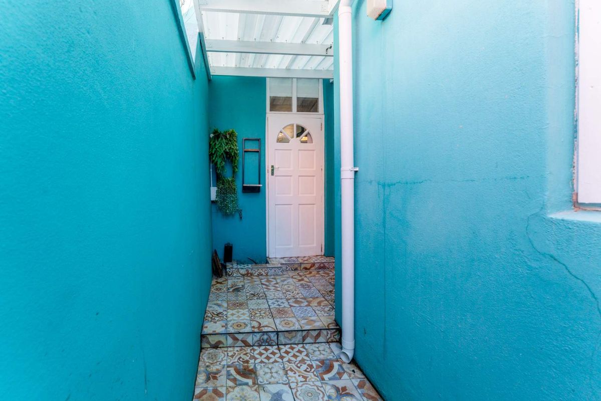 A narrow exterior side entrance featuring vibrant blue walls, patterned floor tiles, and a white panelled door under a covered walkway.