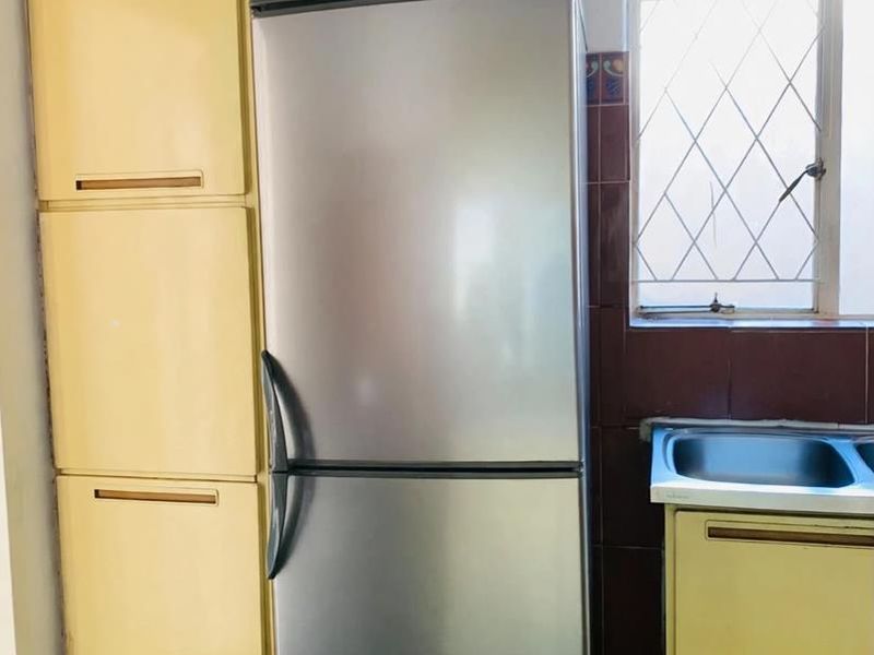 A compact kitchen area featuring dated yellow cabinetry, a stainless steel sink, and dark tiled splashbacks with a window providing natural light.