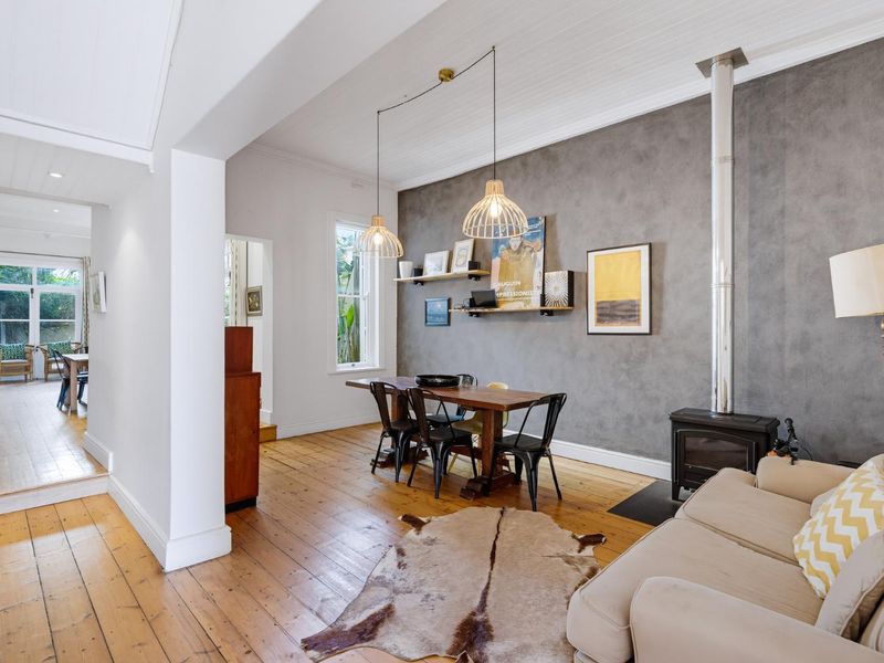 A bright living area featuring polished timber flooring, a feature grey wall with a wood-burning stove, and high beadboard ceilings.