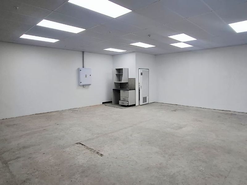 An empty, open-plan office space featuring a suspended ceiling with recessed lighting, concrete flooring, and a small built-in kitchenette unit.