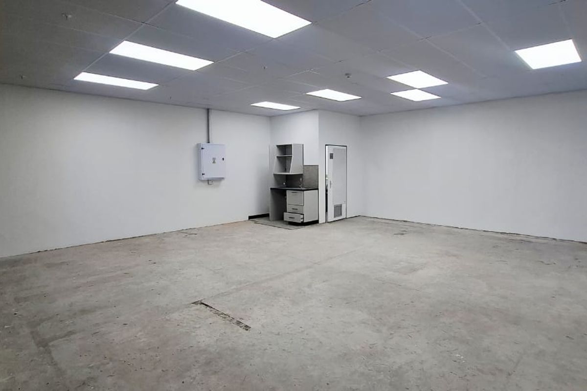 An empty, open-plan office space featuring a suspended ceiling with recessed lighting, concrete flooring, and a small built-in kitchenette unit.