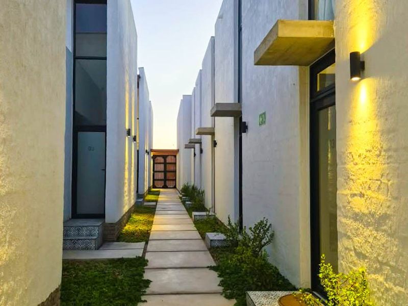 A modern exterior walkway between townhouse units featuring concrete paving, landscaped garden beds, and contemporary wall-mounted lighting.