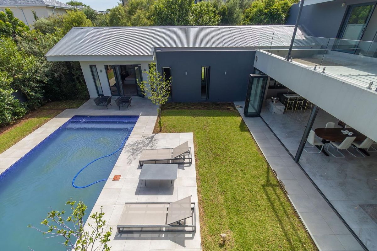 Aerial view of a modern residence featuring a swimming pool, paved patio area, landscaped lawn, and seamless indoor-outdoor flow through large glass sliding doors.