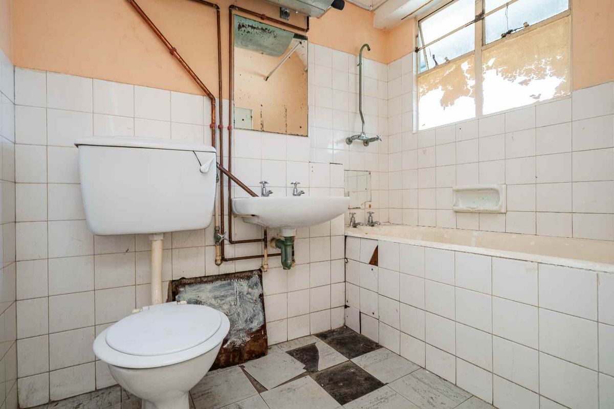 A dated bathroom featuring a toilet, pedestal basin, and bathtub with exposed plumbing, requiring significant renovation and modernization of finishes.