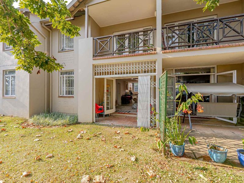 Rear view of the apartment building featuring a ground floor covered patio, an upper-level balcony with wooden railings, and access to a private garden area.