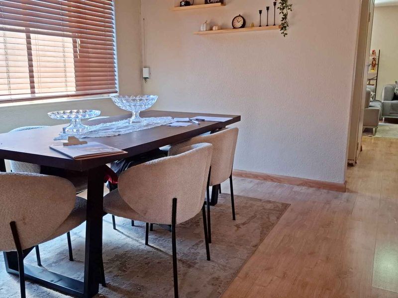 A well-lit dining area featuring laminate flooring, venetian blinds, and wall-mounted shelving, with an open flow into the adjacent living space.