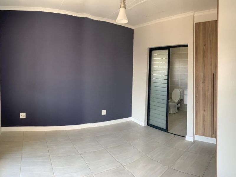 A bright, empty bedroom featuring tiled floors, a dark accent wall, built-in wooden cupboards, and an en-suite bathroom access.
