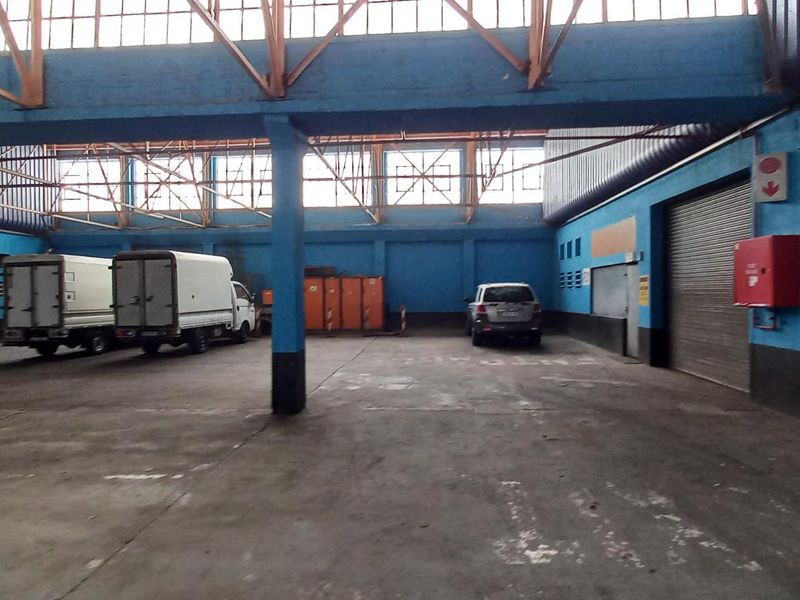 Spacious industrial warehouse or garage area featuring high ceilings with exposed steel trusses, concrete flooring, and a large roller shutter door for vehicle access.
