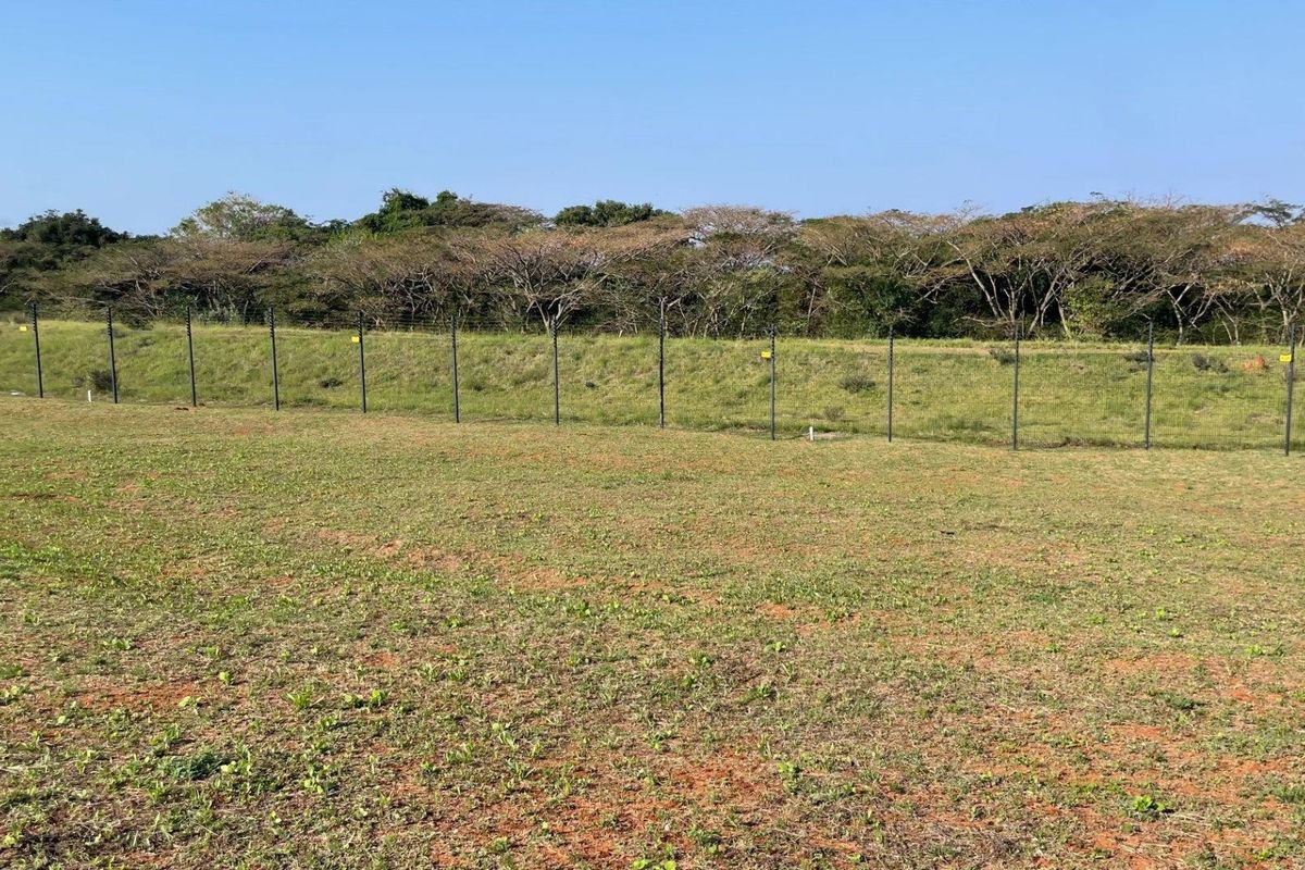 A view of a vacant residential plot featuring cleared grass terrain and perimeter fencing against a backdrop of mature trees.